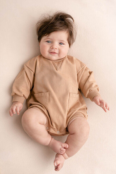 5 month old baby dressed in a khaki sweatshirt romper on a beige backdrop. Baby has dark hair and is smiling up at the camera while lying on back. Portland Portrait Photography.