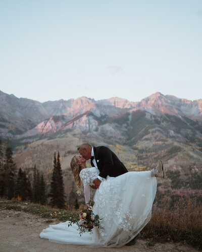 groom and bride are standing next to each other looking out into the distance at an alpine lake in breckenridge. the bride is holding her flowers and she has her hand on the groom's shoulder. there are mountains with snow in the background.