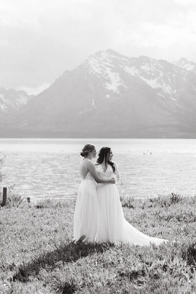two brides embracing next to a lake with mountains in the background