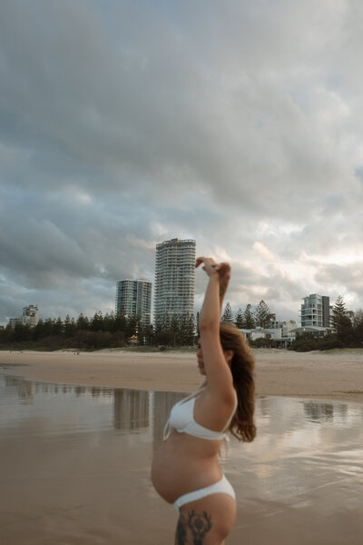 A pregnant mother is dancing in the water reflections of the city scape behind her at Burleigh Beach