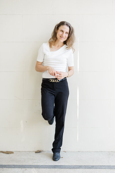 Portrait of Julie-Anne Photography, leaning up against a cream wall posed with one foot lifted and resting on wall behind her.