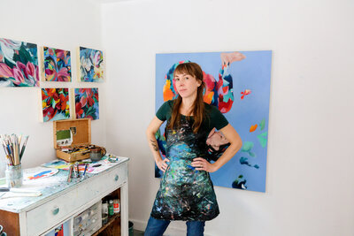 Calgary brand photography of Artist Shantael Sleight standing in her studio with her hands on her hips, her painting and artwork in the background, and her paint brushes on a counter.