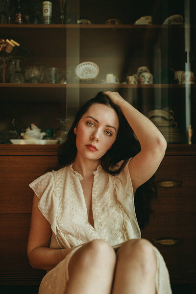 young woman sitting in front of an old china cabinet with her hand in her hair