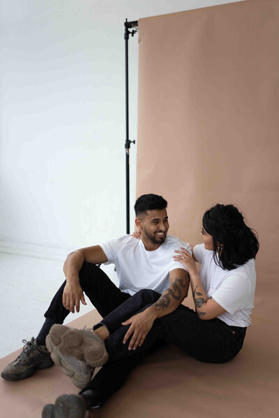 couple sits and looks at each other while cuddling in front of tan studio backdrop