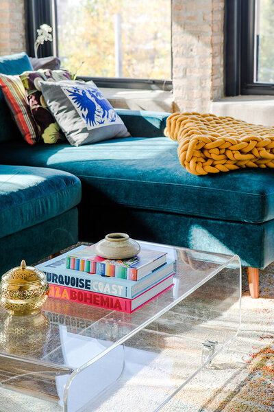 A close-up of a modern living room area. A clear acrylic coffee table holds a stack of colorful books titled "Turquoise Coast," "La Bohemia," and another with a blue and white spine. A small, textured ceramic pot sits on top of the books, and a decorative gold container rests beside them. A plush teal velvet sectional sofa with patterned pillows and a chunky mustard yellow knit blanket is partially visible in the background. Sunlight streams in from a window, casting shadows on the patterned rug underneath the coffee table.