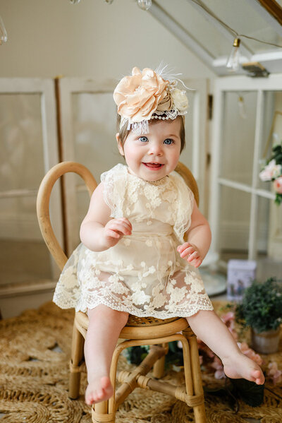 Baby girl in lace outfit and floral headband sits on tiny chair.