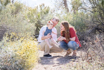 On a bright and sunny morning, a sweet couple crouches to  play with their toddler daughter in a beautiful desert park.
