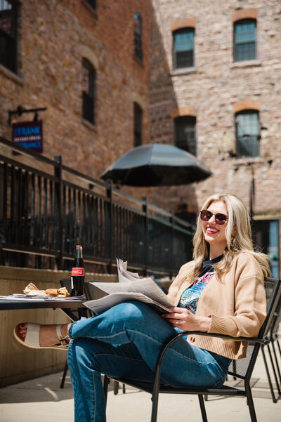 Sarah Klongerbo sitting at an outdoor table wearing sunglasses and reading a newspaper