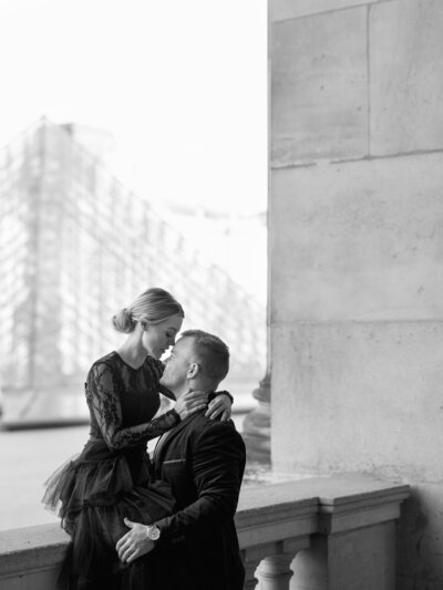 a black and white photo of a woman in a black tulle dress sitting on a  ledge with the louvre pyramid behind her and a man in a suit holding her and looking up at her face