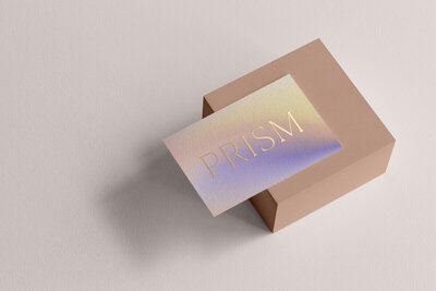 A business card displaying the word "PRISM" in elegant, metallic text is resting on a minimalist, beige rectangular box. The card features a gradient background transitioning from light pink to purple hues. Perfect for showcasing your showit website design, the setup is on a soft, light beige surface.