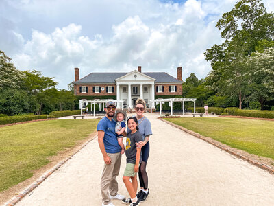 A family of four stands on a path in front of a large, two-story house with white columns and brick exterior. The sky is partly cloudy, and there are trees and greenery on either side of the path.