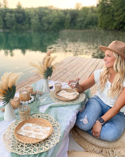 A woman sitting next to a picnic table set up with a blue linen, pampas grass, plates, and napkins.