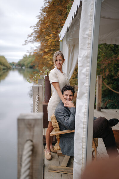 A color photograph of a couple on a boat dock. A man in a blue shirt is seated while a woman in a white dress stands behind him, leaning affectionately. They are framed by a white tent structure with a river and trees in the background.