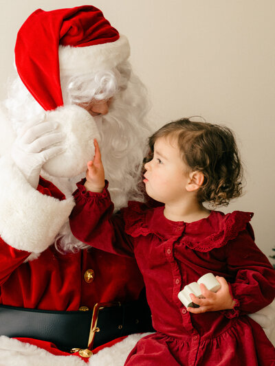 A little girl reaches to feel the fluffy white puff ball on Santa Claus' hat. 