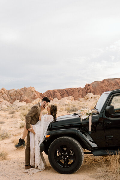 bride is wearing a lacey, boho dress with hiking boots sitting up against a black Jeep in the desert