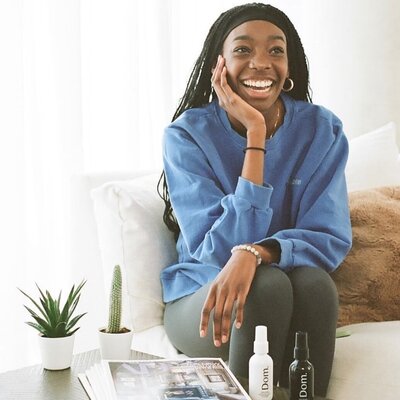 Photo of Tomachi sitting on a white couch, there are skincare products on the table in front of here. She is resting her face on her palm, and smiling past the camera. She is a young black women with braids, and she is waring a blue crewneck sweater.