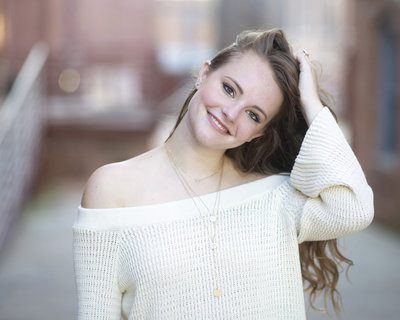 High school senior girl brushes back hair and smiles in white off shoulder sweater