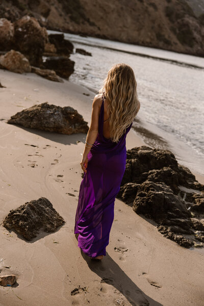 Vrouw in paarse jurk loopt weg van Ibiza fotograaf. Op het strand tussen de rotsen.