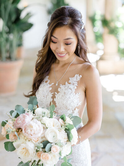 Kohnur Romantic bridal makeup with soft curls, peach blush, and nude-pink lips, photographed with blush and white bouquet in garden venue.