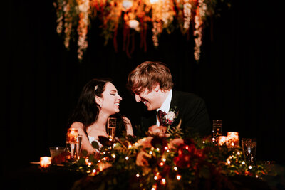 A wedding couple smiling during the reception speeches with a black backdrop and florals in the background and foreground at Registry Bistro in Toledo Ohio 