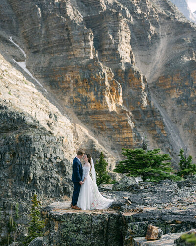 On a mountaintop in Canmore, the bride and groom stand close, exchanging a heartfelt gaze after their adventurous hiking elopement session, surrounded by panoramic mountain views.