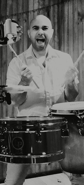 Black-and-white image of a male percussionist mid-performance, holding drumsticks and playing congas with high energy.