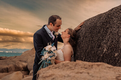 he bride and groom stand on the rocks, embracing as the sun sets, casting a romantic glow around them.