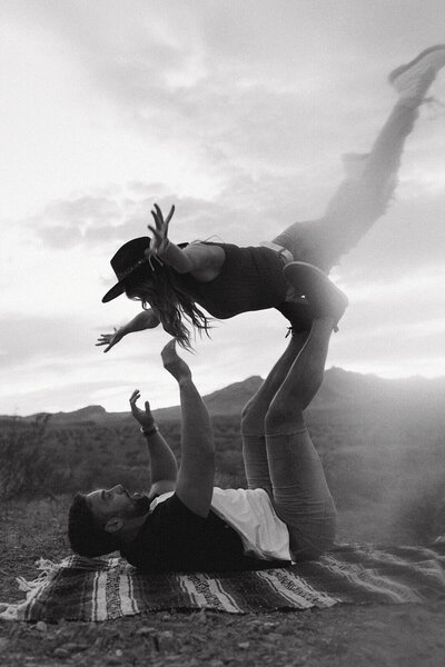 The guy is lying on a blanket with his legs up, holding the girl on top of his legs in the desert