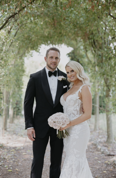 Danielle Armstrong and Tommy Edney on their UK wedding day. Danielle in a Berta dress, Tommy in a tuxedo, framed by a green archway.