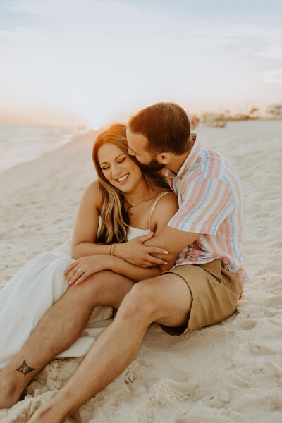 Family Photographer,  a couple embrace as they sit in the sand  at the beach