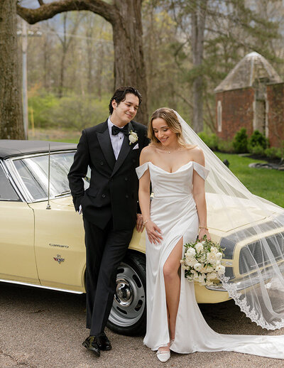 A bride and groom smile together in front of a bright yellow car, capturing a joyful moment on their wedding day.