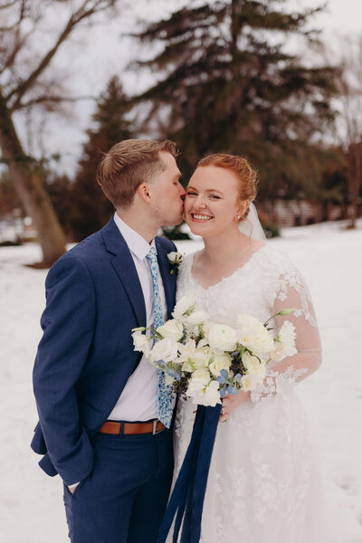 groom kissing bride on cheek surrounded by snow