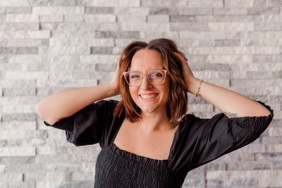 Rachel Hahn, owner, poses against grey brick wall smiling at the camera with her hands in her hair.