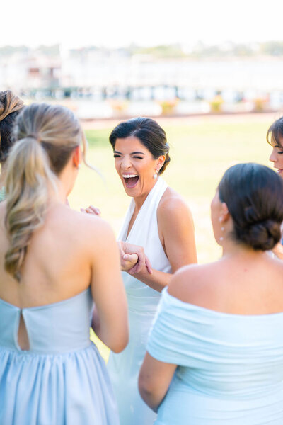 Rhode Island Wedding Photographers | A joyful bride in a white dress laughs with four women in pastel dresses outdoors. 