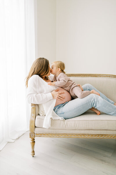 Mom sits on couch in Raleigh photo studio and kisses toddler daughter during maternity session