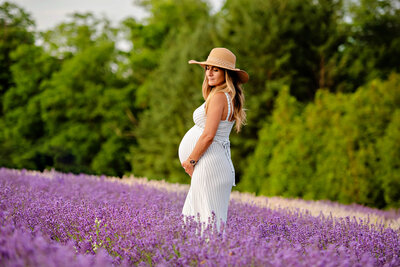 Pregnant lady wearing a striped dress and beige hat standing in the lavender field for her outdoor maternity photos in Burlington, Ontario