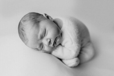 A black and white close-up of a newborn baby peacefully sleeping on a soft surface, curled up in a cozy pose.