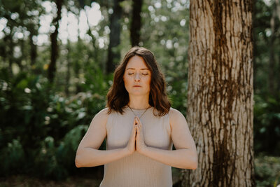 Brandi of The Intuitive Momma stands with eyes closed outdoors and in prayer position. Learn more about her 1:1 Intuitive Sessions.