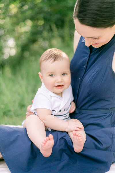 A small child sits on his mother's lap on a picnic blanket - photo by Rochester NY newborn photography service Emi Rose Studio.
