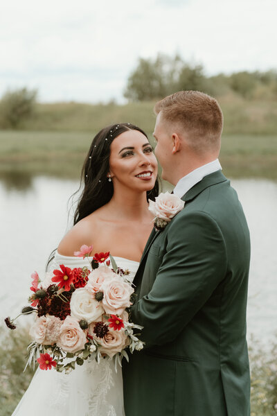 Photo taken by Bailee Freeman Photography of a bride and groom up close, staring in to eachother's eyes/ The groom is wearing an emerald green suit and the bride has dark black hair, green makeup, and a bold bouquet made of red and burgundy and cream flowers. 