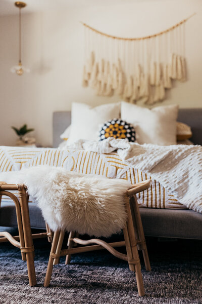 A cozy bedroom with a modern boho style, featuring a gray upholstered bed with layered pillows and striped bedding, a macramé wall hanging, and a wooden stool with a fluffy white throw representing a colorful interior design by Winsome Home Interiors.
