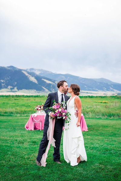 Hot pink and greenery focused wedding  reception tablescape outdoors with bride and groom sharing a kiss at The Big Yellow Barn in Bozeman Montana