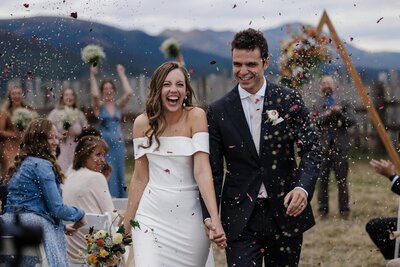 Bride and groom walk out of ceremony while guests toss flower petal confetti.