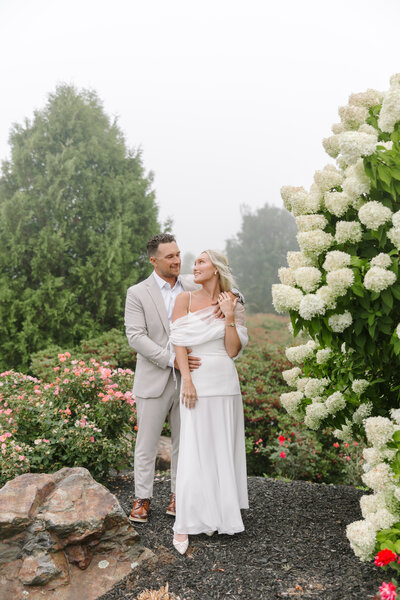 Photo of bride and groom standing outside next to white hydrangea bush while smiling at each other taken by Maine coast wedding photography 