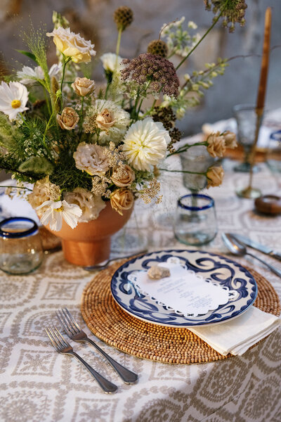 Mexican-inspired wedding tablescape with blue and white Talavera-style plate, neutral florals, woven charger, and custom menu design by Eventive Designs.