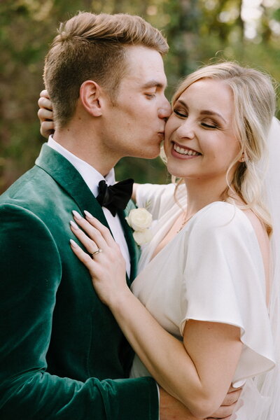 Groom in green suit kisses his bride on the cheek outside in Texas.