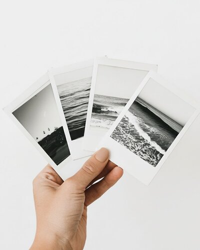 Hand holding four black and white Polaroid photos of the ocean and coastline, styled against a minimal white background.