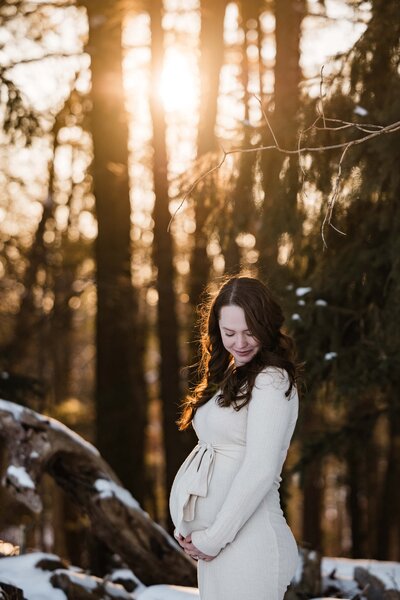 Pregnant woman smiling gently while cradling her belly in a snowy forest with sunlight filtering through the trees, captured by a talented maternity photographer from Pittsburgh.