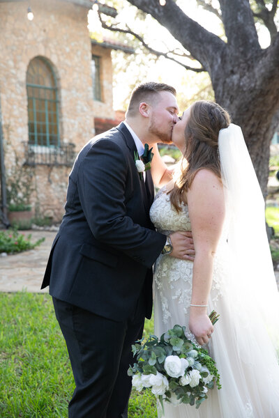 An Austin-based wedding photographer capturing a heartfelt moment as the bride and groom share a sweet kiss in front of a beautiful tree.