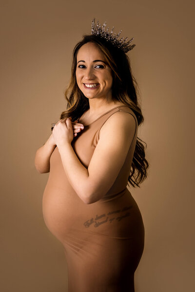 Radiant pregnant woman wearing decorative crown smiles joyfully in sheer tan gown during glamorous studio maternity session captured by Connecticut maternity photographer